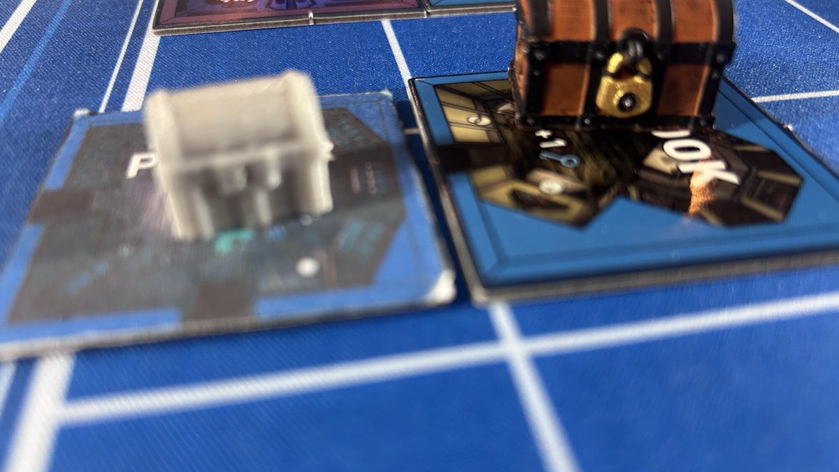A small gray unpainted chest miniature beside a large painted treasure chest on the mat