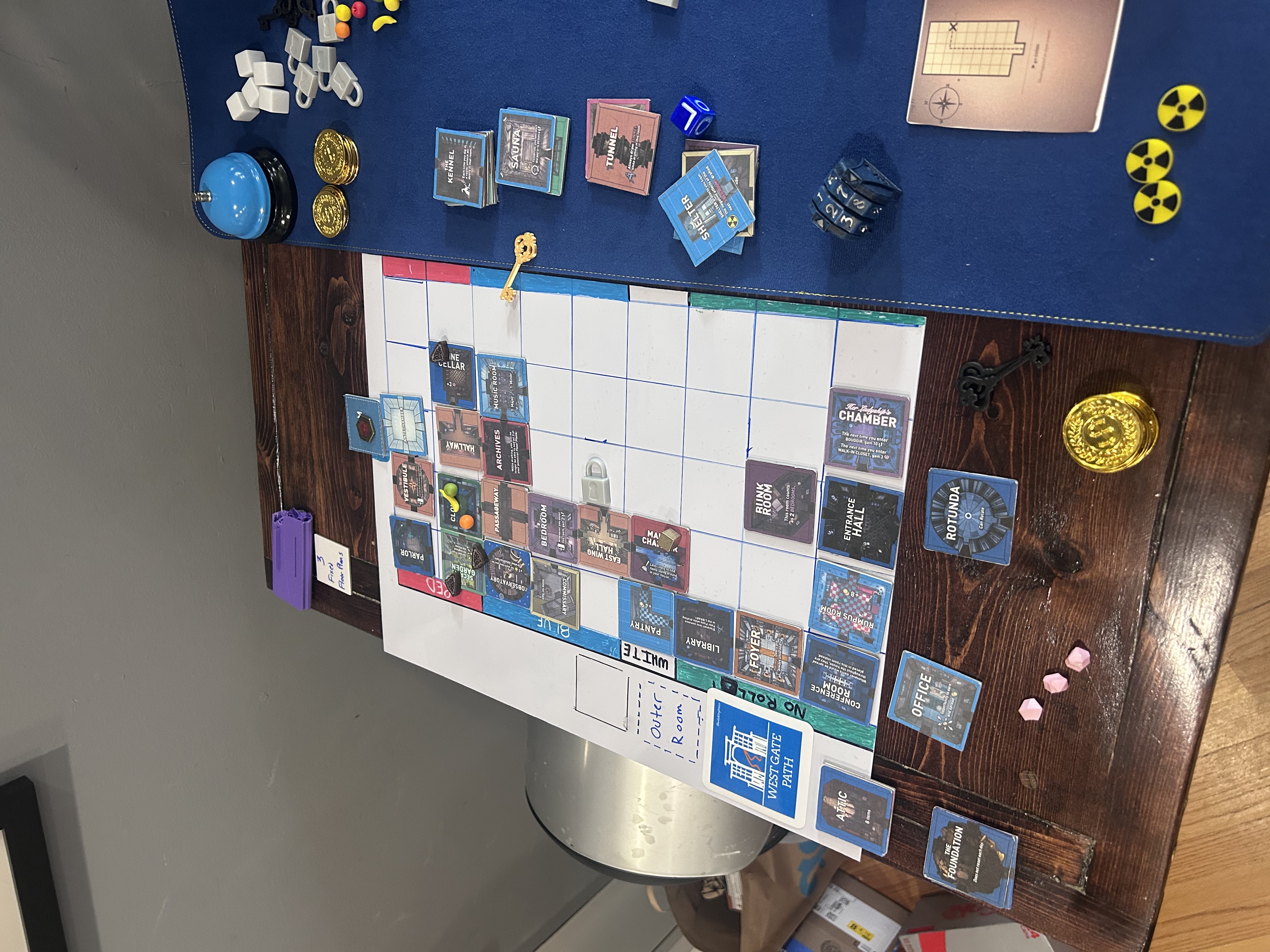 Side angle of the board mid-game — rooms placed in a winding path from Entrance Hall toward the upper ranks, outer room stacks visible on the mat to the right, gold key on the board
