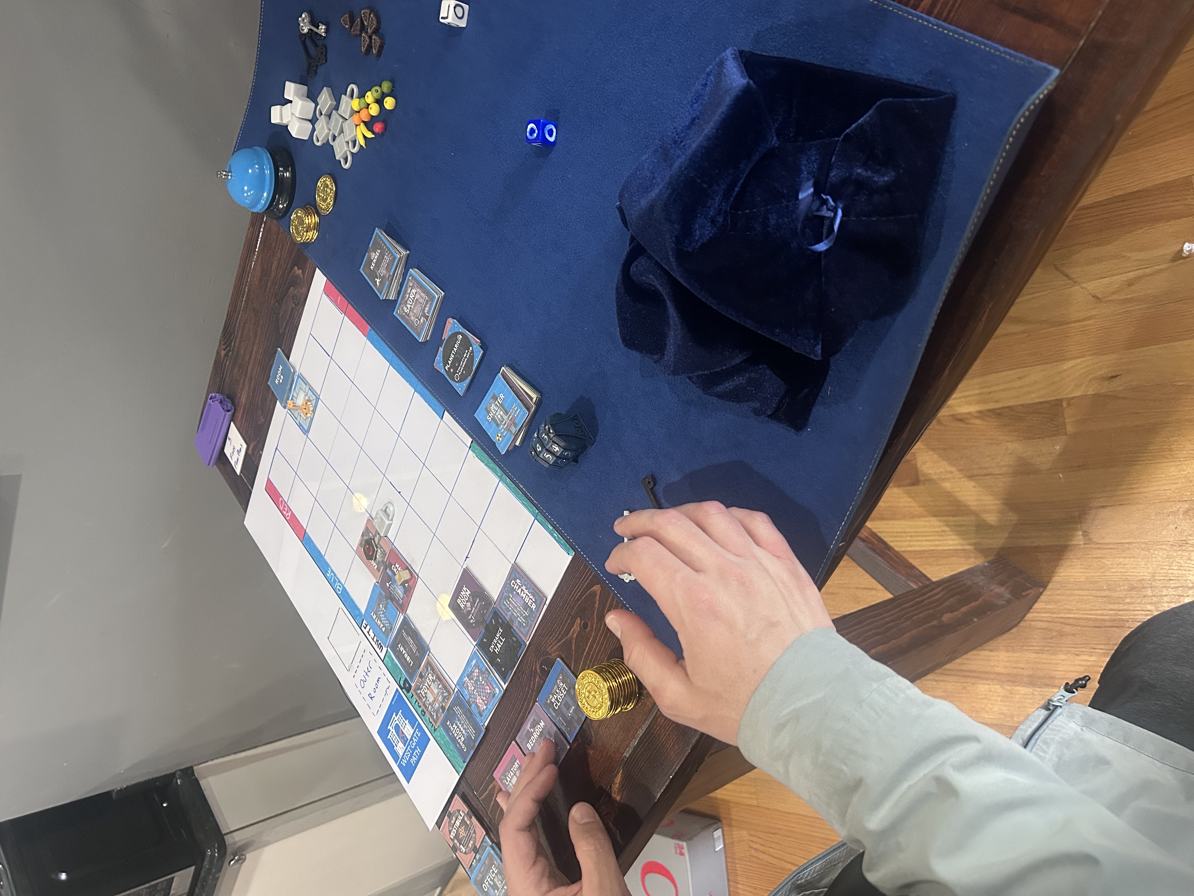 Wide shot of the Blue Prince board game prototype in progress — Mike's hands reach across the navy mat toward the paper board, tokens and the velvet tile bag visible