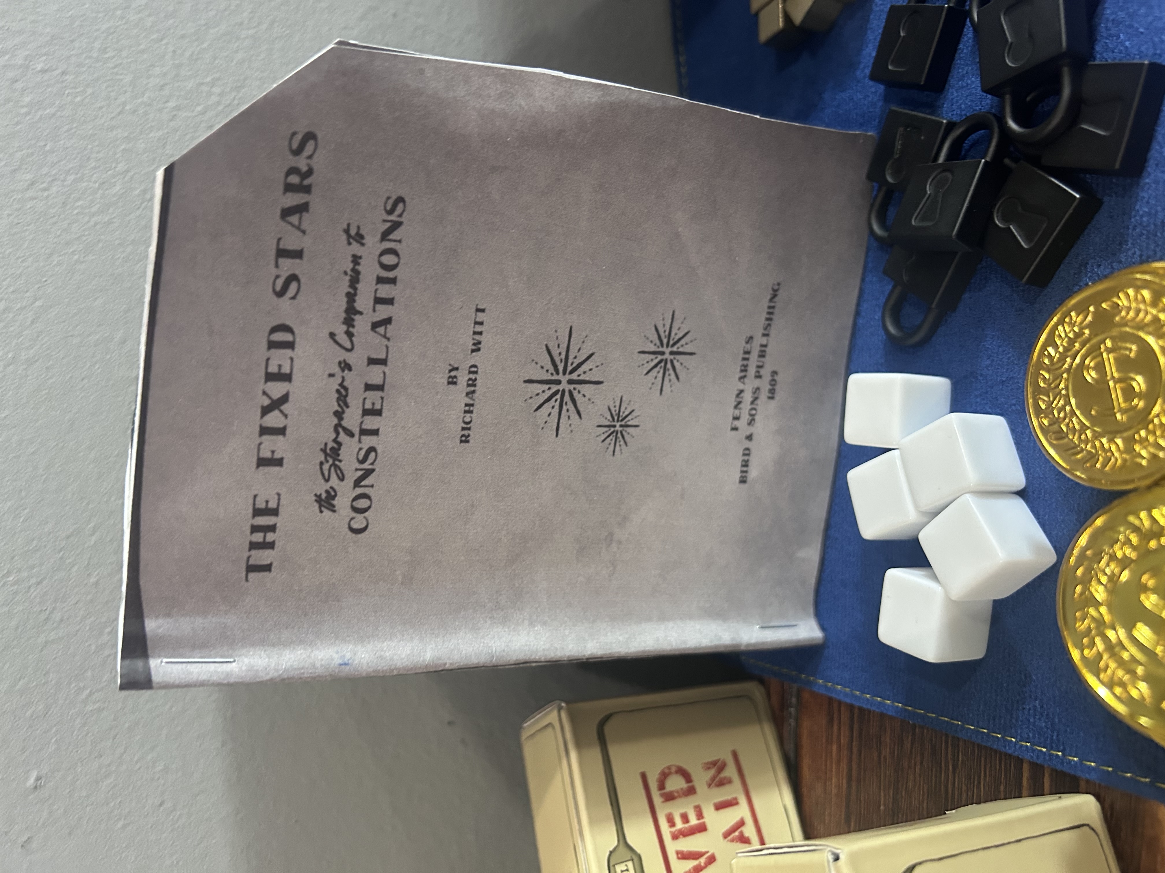 The Fixed Stars booklet standing upright on the navy mat, white blank dice and black lock tokens visible in the foreground alongside gold coins