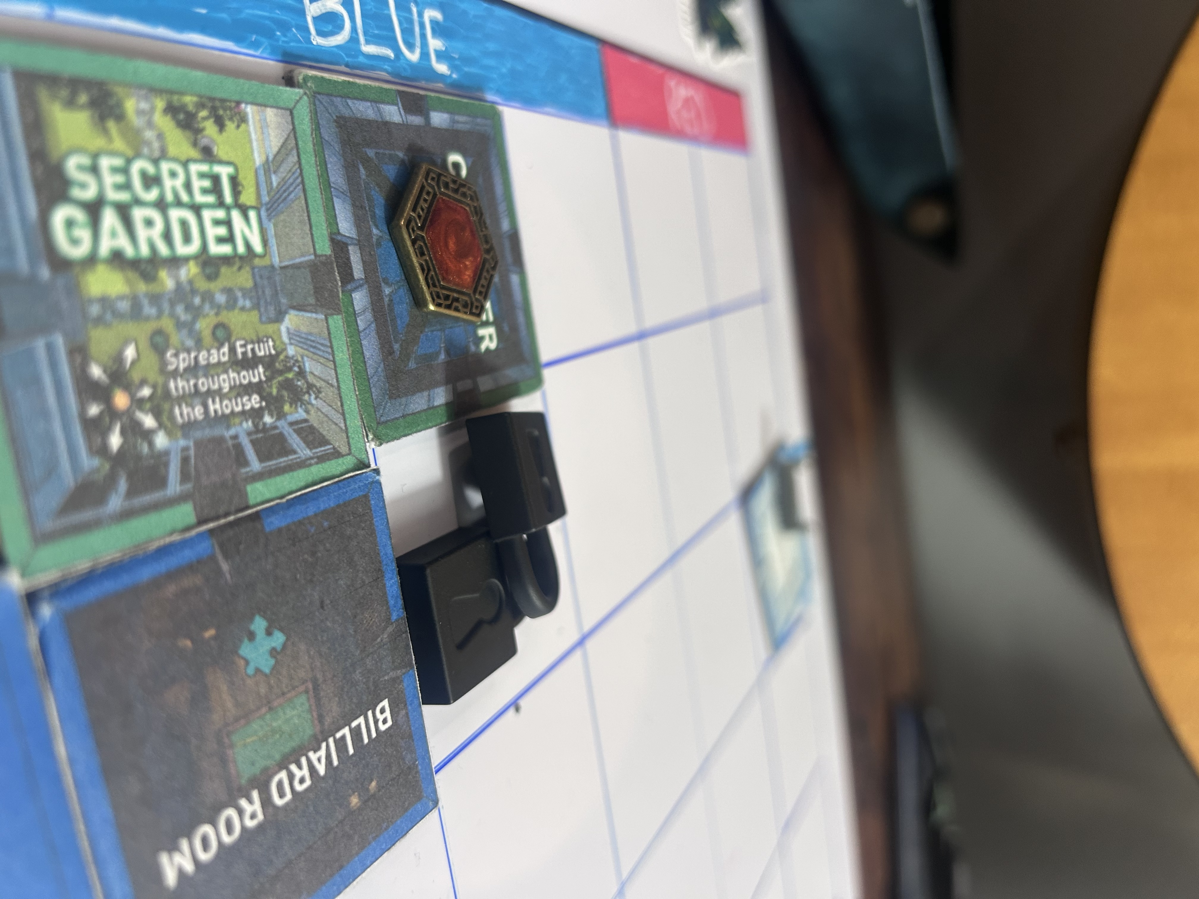 Close-up of two black lock tokens stacked and overlapping on the Cloister tile corner, Secret Garden tile below, Billiard Room to the right, blue L die visible above