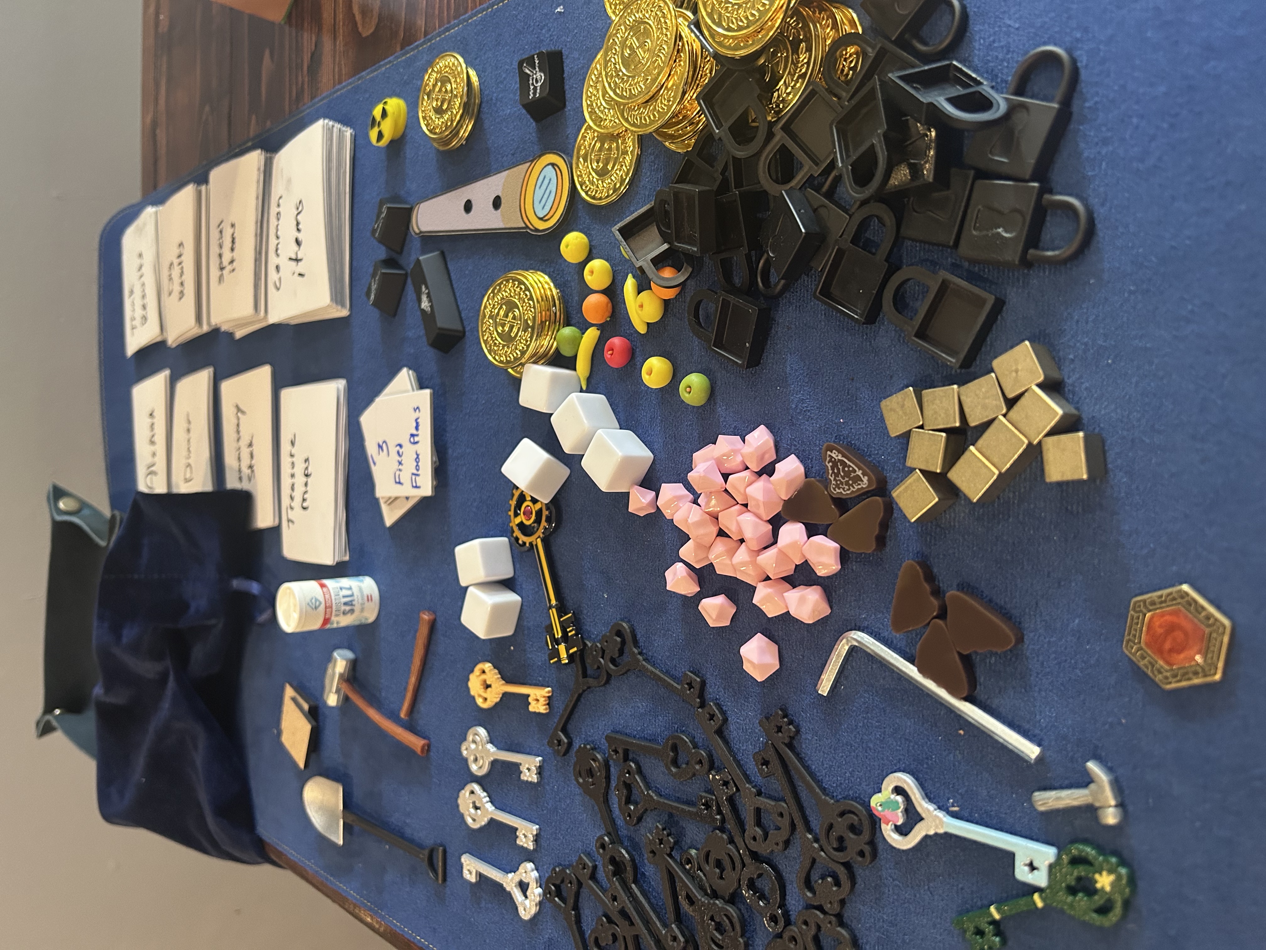 All game components laid out on blue velvet — keys, gems, padlocks, coins, food tokens, cards, brass cubes