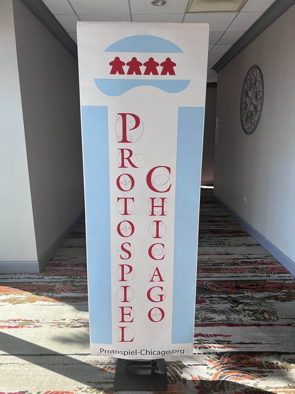 A tall Protospiel Chicago banner standing in a hotel hallway, red lettering on white with four red meeples across the top