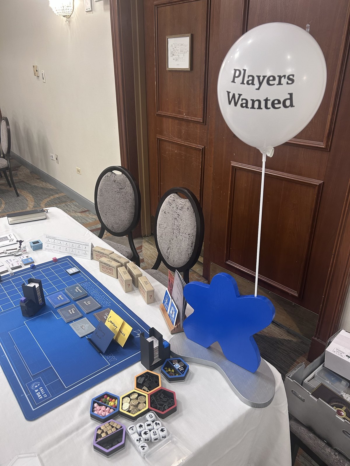 The playtest table at Protospiel: the blueprint mat with two dealer towers, card decks, Archived Keychain boxes, sticky-note labels, trays of tokens, two empty chairs, and a white balloon reading Players Wanted