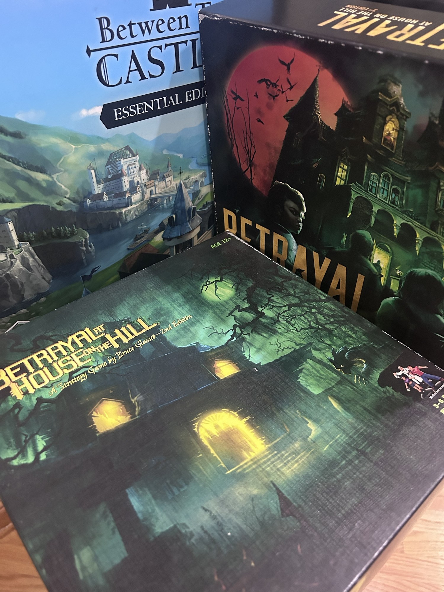 Three board game boxes stacked on a table: Betrayal at House on the Hill 2nd edition in front, the 3rd edition behind it, and Between Two Castles Essential Edition at the back