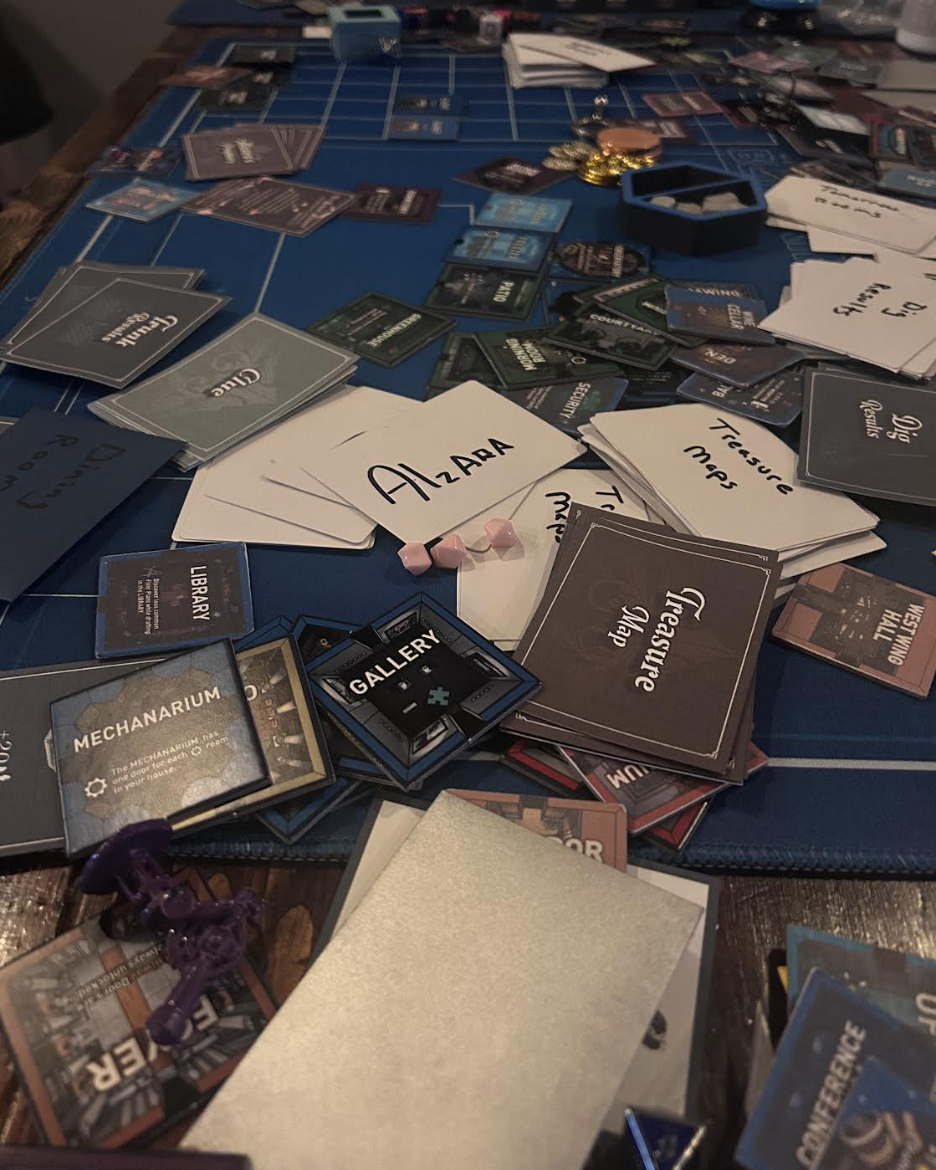 A cluttered tabletop covered in hand-labeled sorting index cards, finished deck backs, room tiles, tokens, and a painted telescope miniature, mid-organization