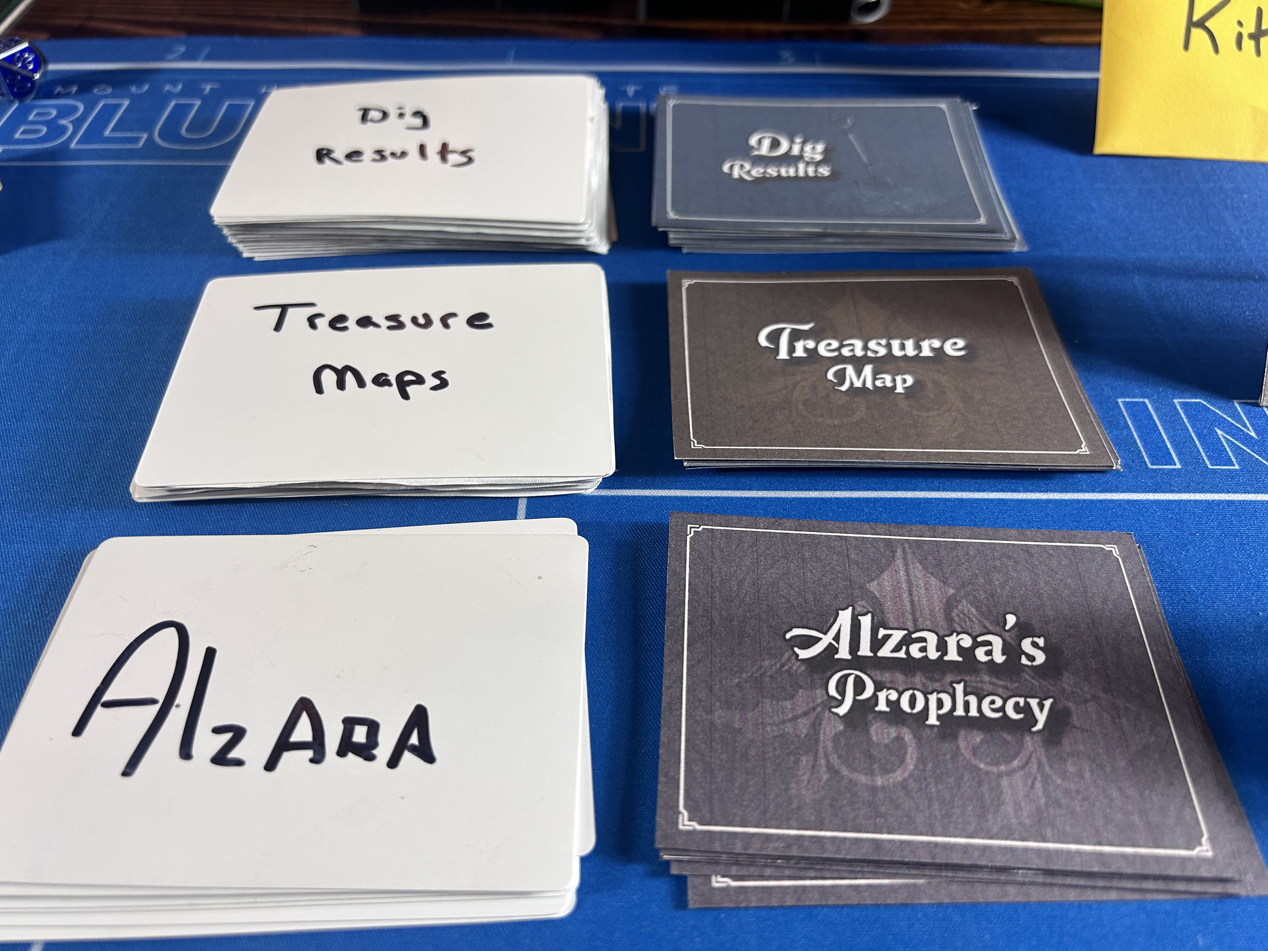 Six stacks of cards on the blue board, each with a hand-labeled paper marker: Dig Results, Treasure Maps, and Alzara's Prophecy visible with their new printed backs