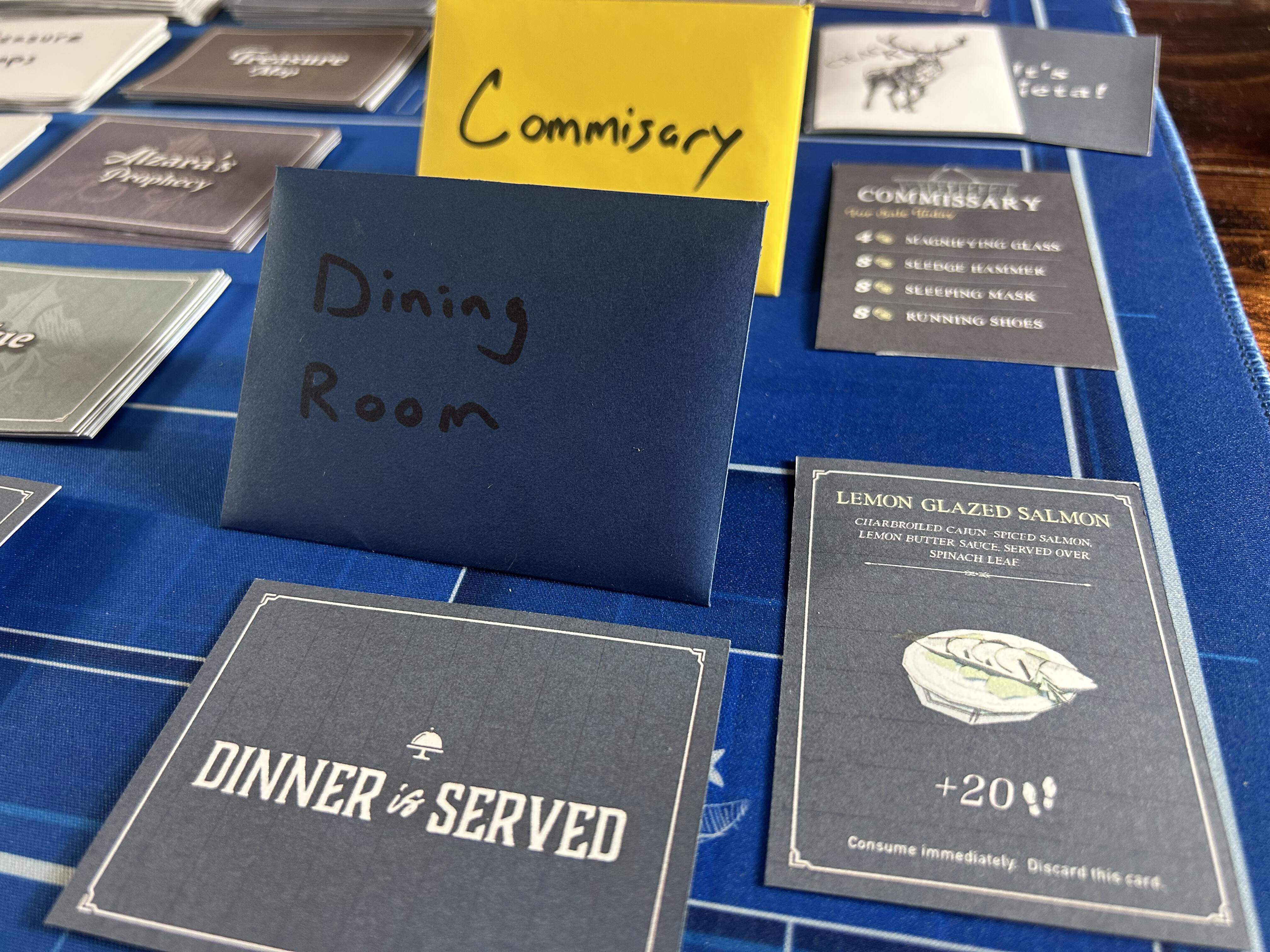 A Commissary shop card listing priced items, a Dinner is Served card, and a Lemon Glazed Salmon meal card with illustration and flavor text, next to a navy envelope labeled Dining Room by hand