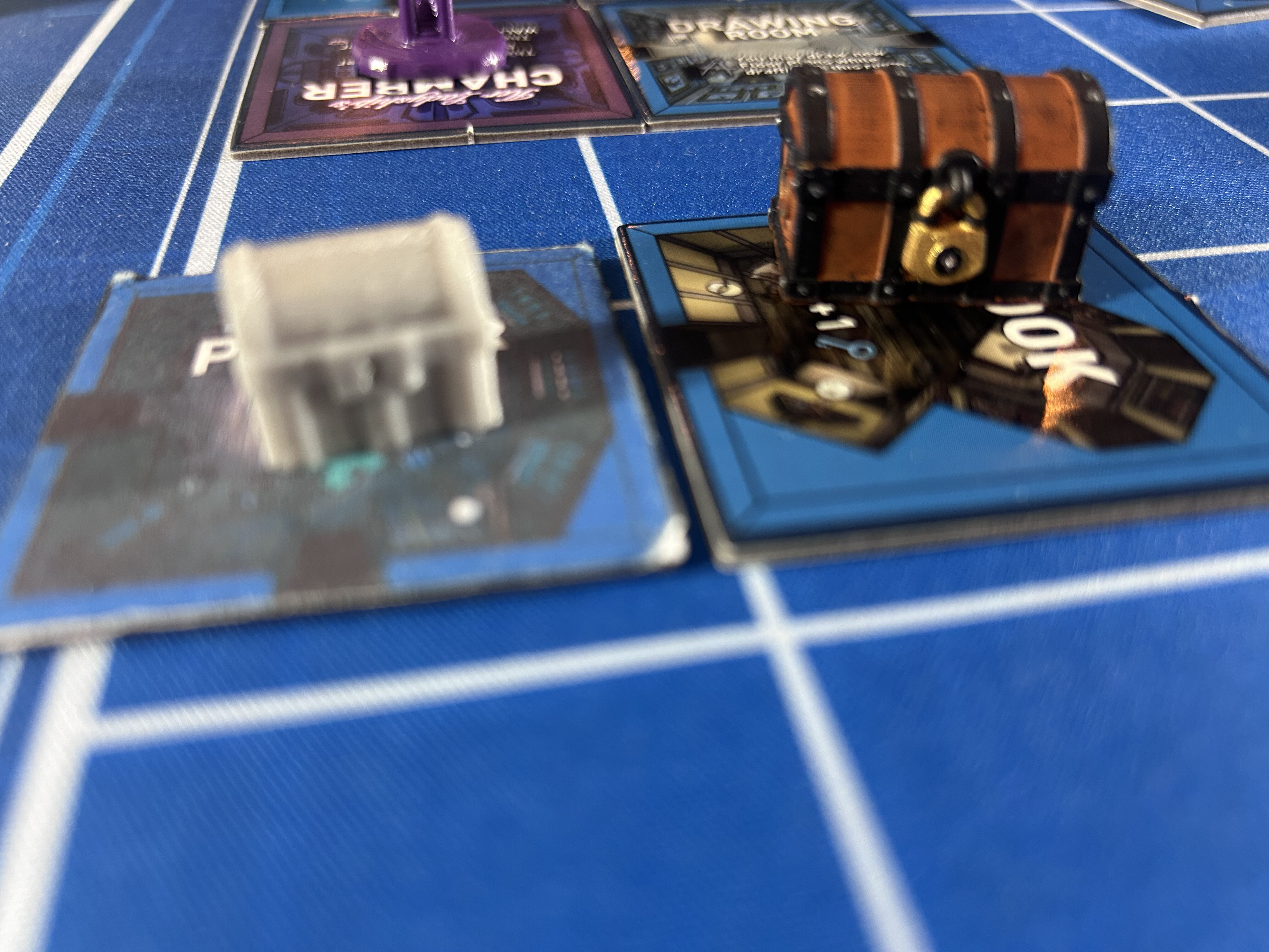 An unpainted gray resin miniature beside a fully painted treasure chest miniature, sitting on room tiles on the blue Mount Holly Estate board